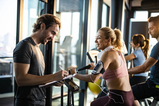 gettyimages-1212327852-612×612 Happy woman exercising on stationary bike in a gym and looking at her coach making plans for her future sports trainings.