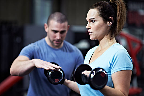 gettyimages-470333831-612×612 Healthy female lifting dumbbell free weights in gym with encouragement from personal trainer