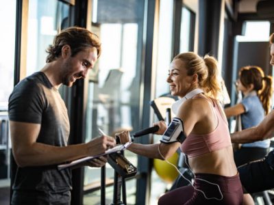 Happy woman exercising on stationary bike in a gym and looking at her coach making plans for her future sports trainings.