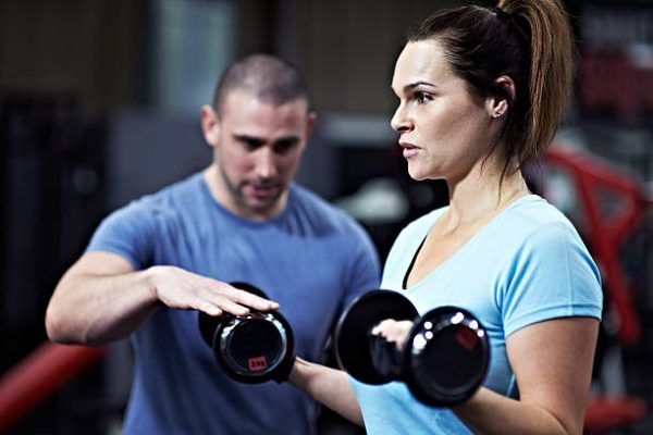 Healthy female lifting dumbbell free weights in gym with encouragement from personal trainer